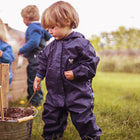 A child wearing the 'Originals Waterproof Puddle Suit Navy' playing with a stick in a bucket outdoors on a grassy field.